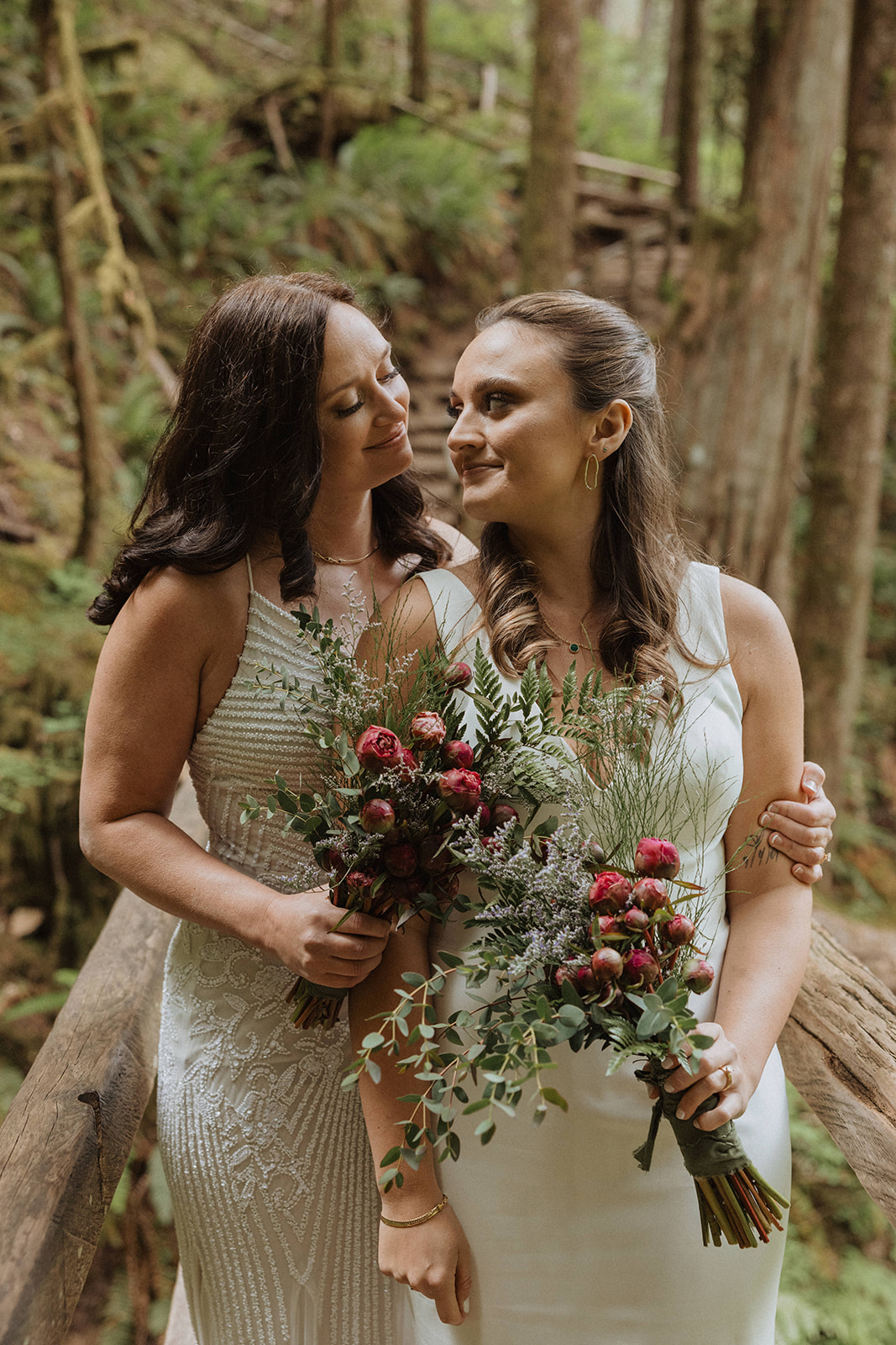 Female couple on their wedding day, both in white dresses, carrying bouquets, in the woods at Olympic National Park