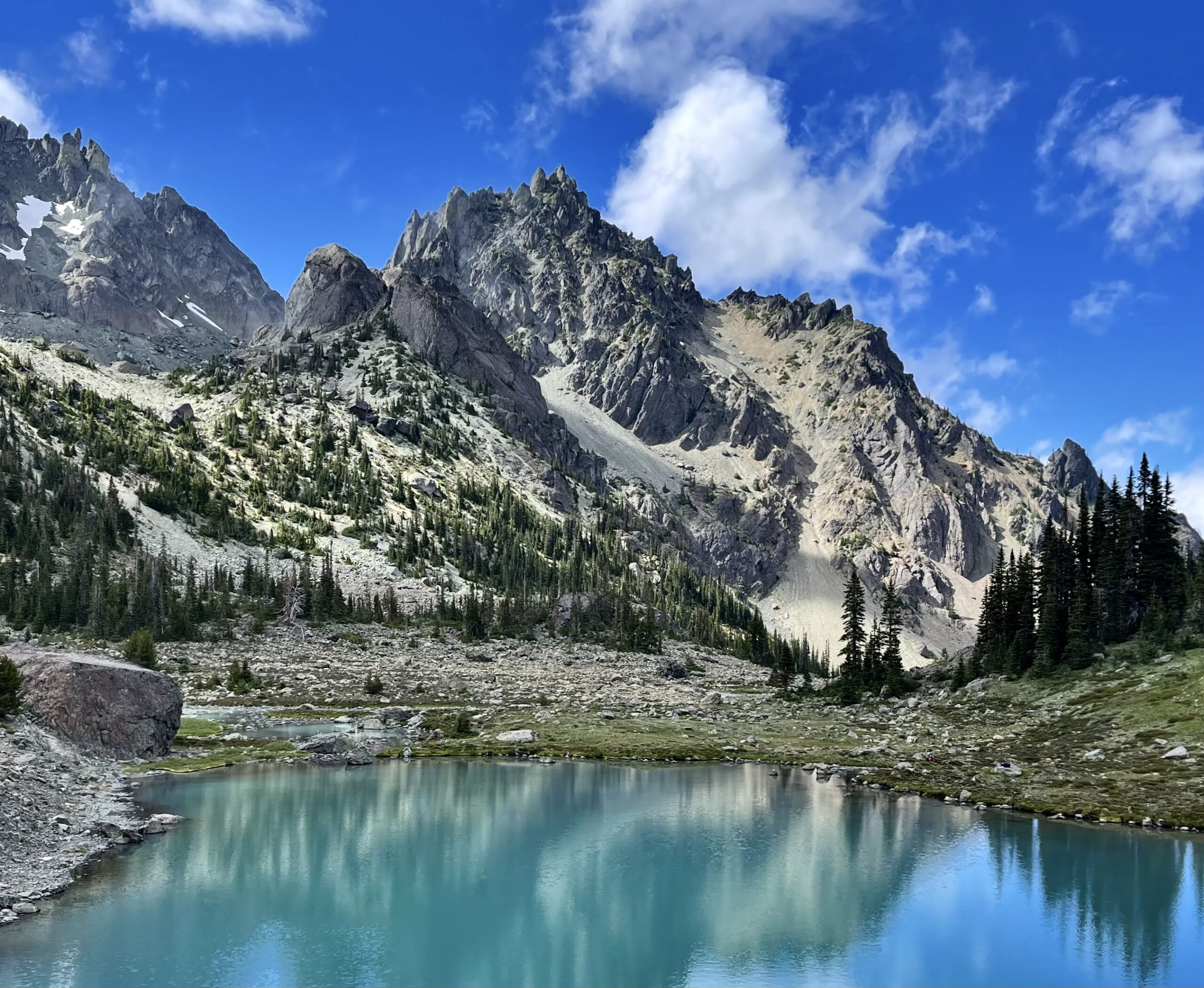 Pictured is an aqua blue lake in the foreground with trees behind, and snow-capped mountains behind that, all on a field of a bright blue sky with wispy white clouds